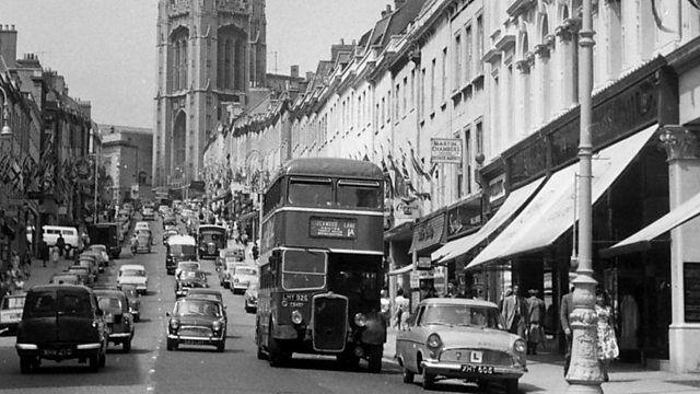 A black and white image of a bus coming down Park Street in Bristol in the 1960s. There are cars coming up and down the road, and the Wills Memorial Building can be seen in the background at the top of the hill.