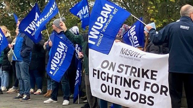 About a dozen protesters carrying blue NASUWT banners. There is also a larger white banner being held by people on the right of the photo. It reads "On strike fighting for our jobs".