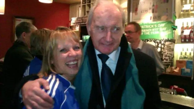 A picture of a woman in a Chelsea shirt next to an elderly Alan Hudson who has her arm around her shoulders
