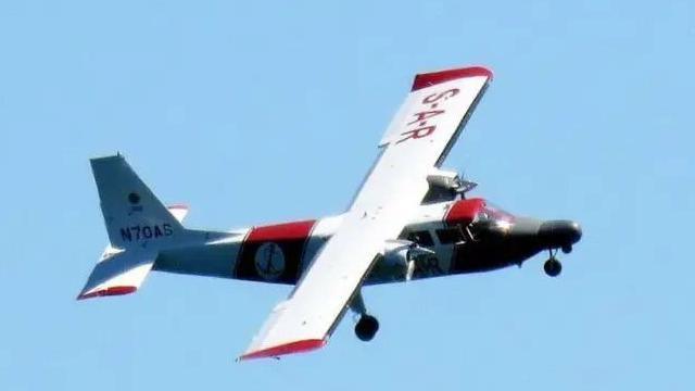 The aircraft, called the Lions’ Pride, airborne. It is white with red writing on the right wing which says 'S-A-R' and has a red block in the middle. The wing at the back has red text on it. The the front there are two windows on the left and is grey.