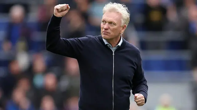 David Moyes, wearing a black top and looking serious, clenches his first towards supporters. A blurred crowd is in the background.