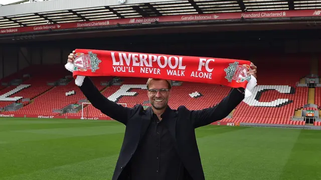 A smiling Jurgen Klopp, wearing a black suit and shirt, holds a Liverpool scarf above head on the pitch at Anfield with the empty Kop behind him after joining in October 2015