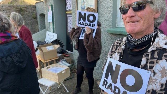 Campaigners stand outside a building wearing banners saying "NO RADAR"