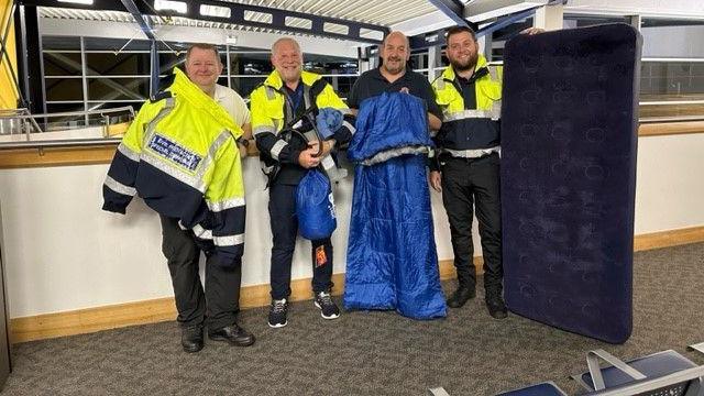 Four civil protection officers wearing high-vis uniforms smile at the camera holding sleeping bags and an inflated mattress in the airport.