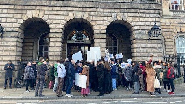 A large group of people stood outside Liverpool Town Hall with placards. The Town Hall is grey brick building with three arches at the entrance.