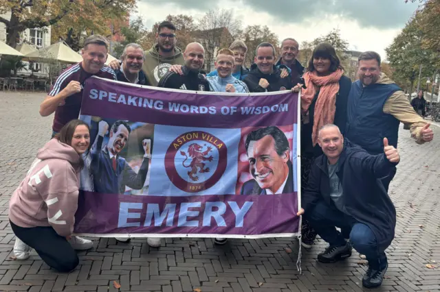 A group of Aston Villa fans in Deventer holding a flag with two pictures of Unai Emery and the club badge on it, with the text: "Speaking words of wisdom, Emery"