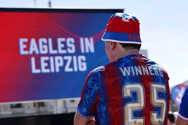 A Crystal Palace fan wears a shirt which says 'winners 25' on the back. In front of him is a big screen which reads 'Eagles in Leipzig'