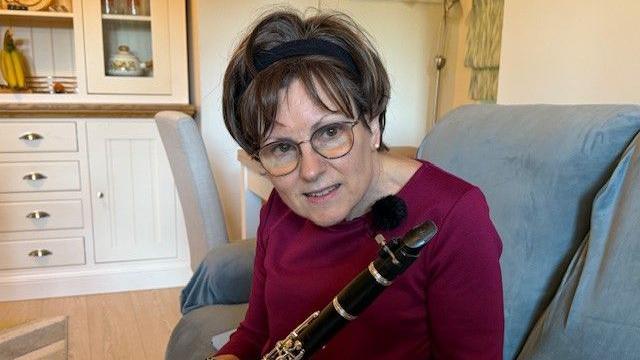 Denise Bacon sits on a sofa holding her clarinet, while wearing a purple top.
