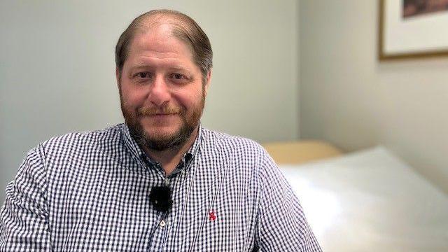 A man in a checked shirt looks at the camera. He is sitting in a plain room with little furnishing.