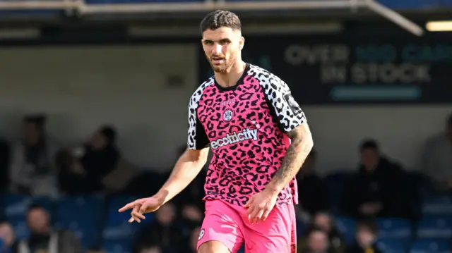 Ryan Inniss running on the pitch, wearing Forest Green's pink away kit, with his right arm out at his side and index finger extended