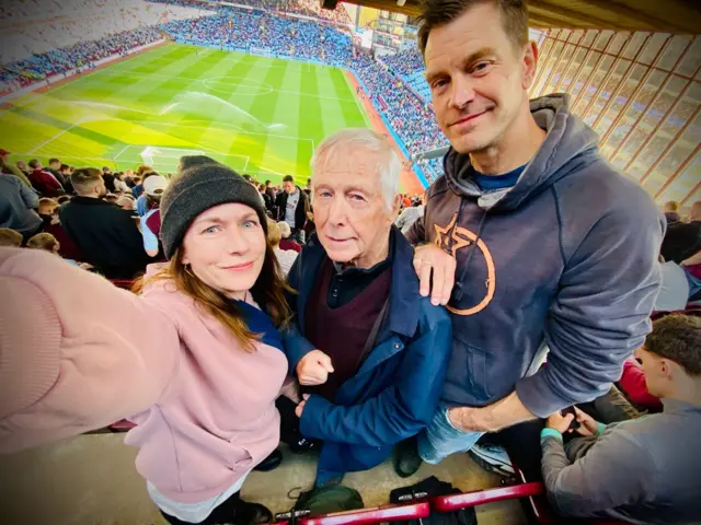 A wide angled picture of a man, a woman and an older gentleman with Villa Park in the background