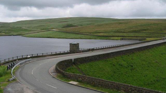 Huddersfield Road snakes above the Dowry reservoir dam, with a basin of water seen behind the route, with green hills in the distances on an overcast day.