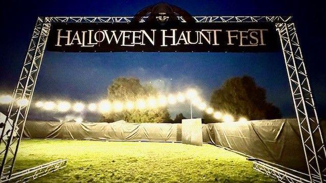 A black entrance sign saying "Halloween Haunt Fest" hanging from a large metal archway. It is on a field with barriers around, draped in white tarpaulin. A string of lights runs across the site and there are trees in the background. No people in shot