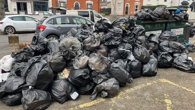 Piles of black bin bags full of rubbish sit on the ground next to overflowing large green bins, along the side of a road with cars parked nearby.