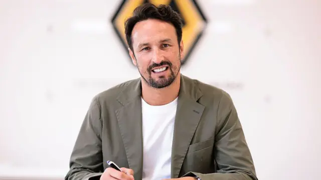 A bearded Wolves boss Cesar Peixoto wearing a grey suit jacket and white t-shirt smiling while signing his contract