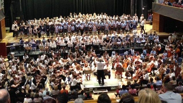 A man in a white shirt is stood in front of a large orchestra and a group of what appears to be school children who are singing at a concert. The photo has been taken from the audience's point of view.