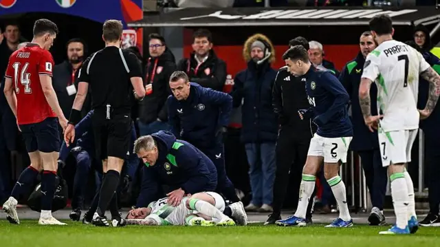 Sam Szmodics of Republic of Ireland is helped by Republic of Ireland head coach Heimir Hallgrimsson after a head injury. Szmodics is wearing an all white Republic kit and laying on the ground