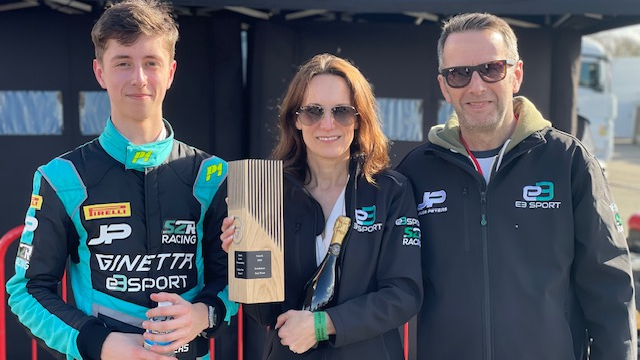 Jude in blue and black race suit, next to his mum, who is holding a bottle of champagne and a trophy, and his dad to her right. Both parents have a jacket promoting Jude's racing team.