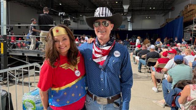 Maleesa Meyers, left, is wearing a red, white and blue Wonder Woman costume and Michael Milton, right, is wearing a black cowboy hat with Trump's name on the front and an American flag scarf along with a button-down long sleeve shift and thick leather belt. The two are photographed at the back of a JD Vance rally in an airplane hangar. 