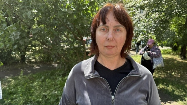 A woman in a grey jacet stands on some grass with a tree behind her.