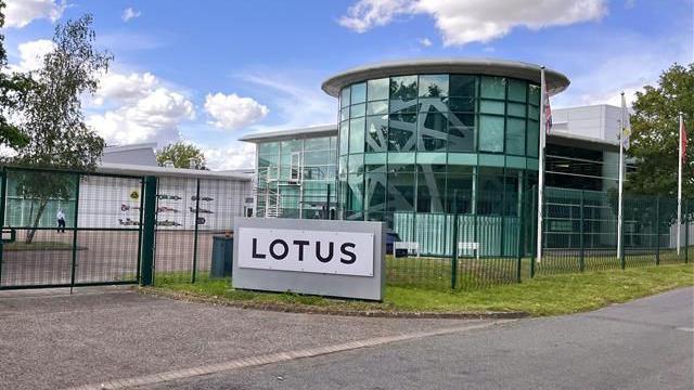 The outside of the Lotus building in Hethel, near Wymondham. The building is a curved glass building with a sign outside the front which says LOTUS in black letters.