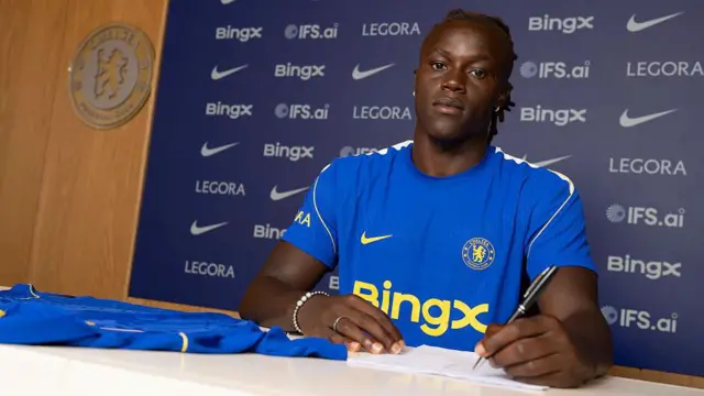 Geovany Quenda signing his contract at a desk with a Chelsea shirt in front of him
