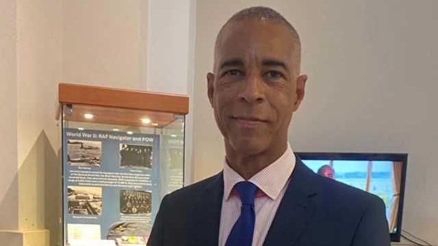Eddy Smythe is smiling at the camera wearing a navy suit, pink shirt and blue tie. In the background is the Thame Museum.