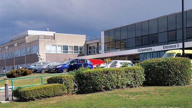 A line of bushes in front of a car park outside a building on the Wythenshawe Hospital building on an overcast day. 