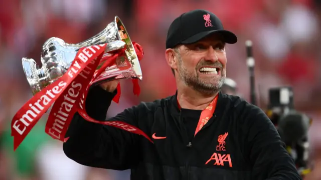 Jurgen Klopp, wearing a dark Liverpool cap and top, grits his teeth as he holds the FA Cup in his right hand