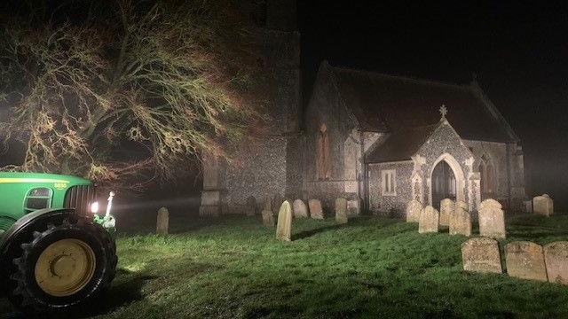 A green tractor shines its light through the darkness on the entrance of a church. The gravestones in the churchyard with green grass are also visible. There is tree behind the tractor.