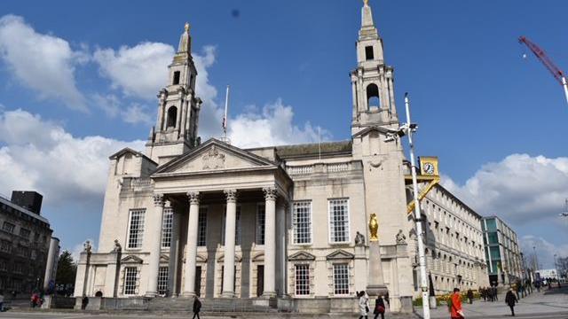 Leeds Civic Hall, a white stone building with a blue sky behind. To the right is a golden clock attached to the building and a golden owl on a pillar. There are pedestrians walking on the pavements outside. 