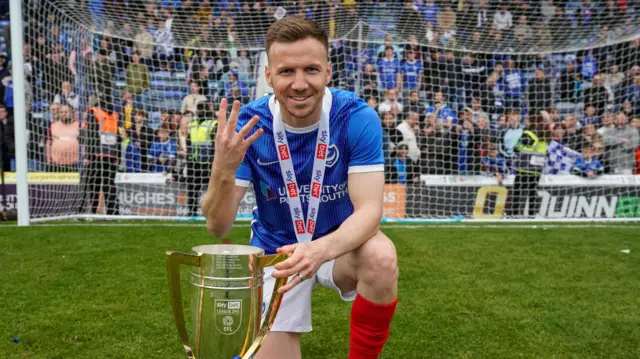 Lee Evans, in a Portsmouth kit, celebrating with the League One trophy which he has his left hand on while kneeling down on his right knee and holding up four fingers with his right hand