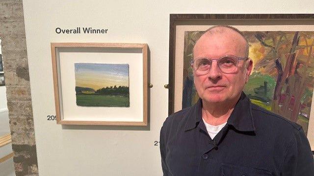 Artist Robert Newton standing next to his painting. Mr Newton has thinning hair and wears clear-rimmed glasses and a dark shirt. His painting shows a green landscape with fields and trees under a blue and yellow sky.