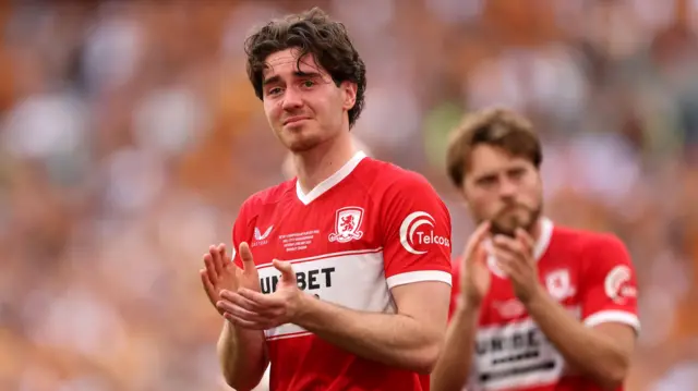 A photo of Hayden Hackney, wearing Middlesbrough's red and white home kit, applauding the fans while looking emotional after their play-off final loss to Hull City in May 2026