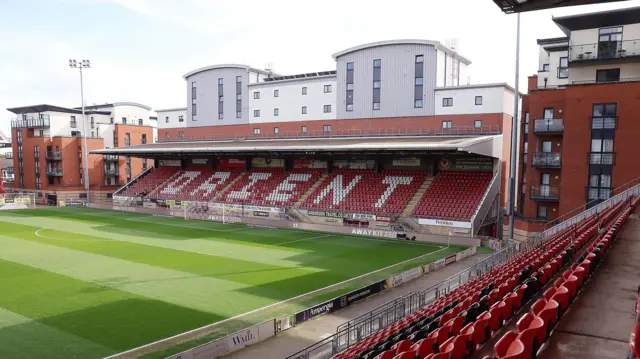 A view from the side of Leyton Orient's stadium, a football stand with red seats with Orient painted in white .