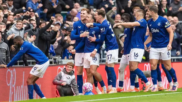 Everton players, wearing blue shirts and white shorts, celebrate their late winner against Crystal Palace, with score Jack Grealish being hugged by team-mates as they all smile at the crowd in the background