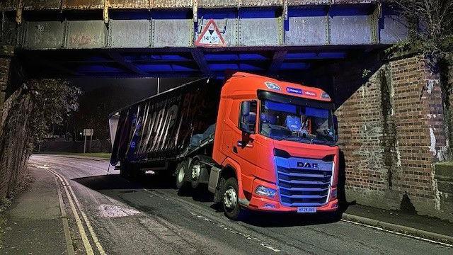 A large lorry with an orange cab and a dark blue trailer is wedged underneath a railway bridge. 
