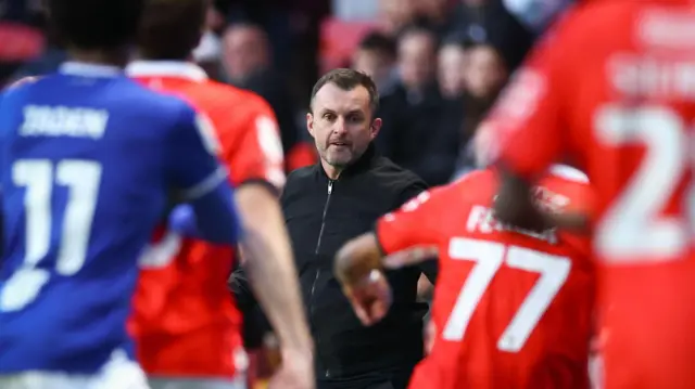 A photo of Nathan Jones in the middle of Charlton and Ipswich players playing football as he watches on while wearing a black zip up jacket