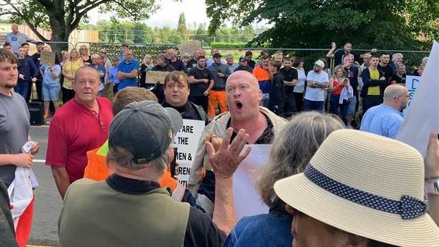 A bald man holding a placard is shouting at a man who has his back to the camera. There are lots of other people around, some holding cardboard signs, across the road