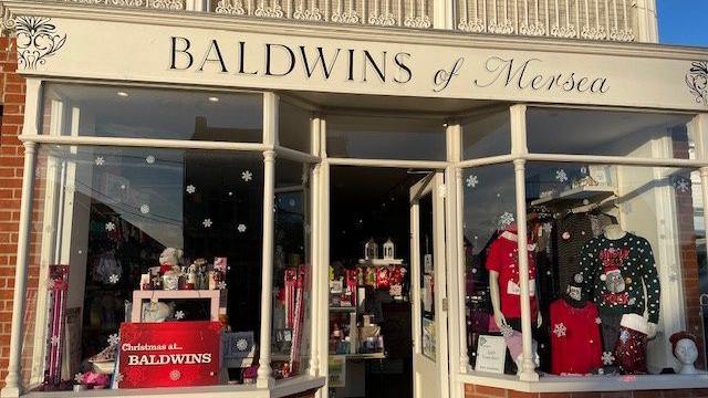 A photo of the outside of Baldwins' Mersea store. It has a white sign which reads Baldwins of Mersea. Christmas clothing and decorations are in the window displays.