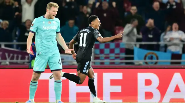 Ollie Watkins, wearing Aston Villa's black away kit, celebrates after scoring as a disappointed Bologna player, in his side's light blue kit, looks angry