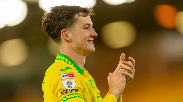 A photo of Kellen Fisher smiling as he applauds wearing his Norwich City home kit
