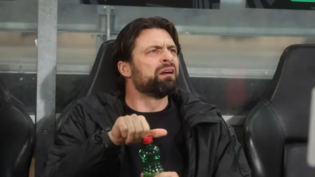 Russell Martin, sitting in the visitors' dugout at SK Sturm Graz while managing Rangers in 2025, looks confused as he opens a green plastic bottle with a red lid