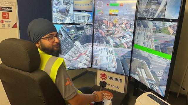 A man with a beard wearing glasses and a turban is sat on a chair looking off to the right. Behind him is a large monitor that has an image of a building site on it