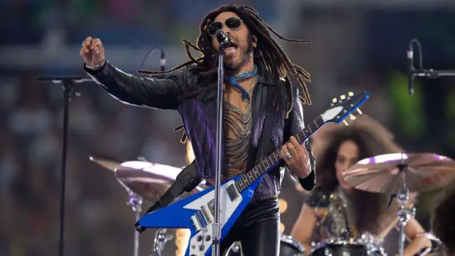 Lenny Kravitz, holding his guitar and clenching his fist, singing passionately during a concert