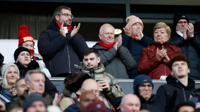 People watching Bristol City with owner Steve Lansdown in the middle applauding wearing a red scarf and a grey puffer jacket