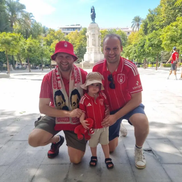 A young boy poses for a photo with his father and uncle in front of a statue in Seville. They are all dressed in Nottingham Forest shirts.