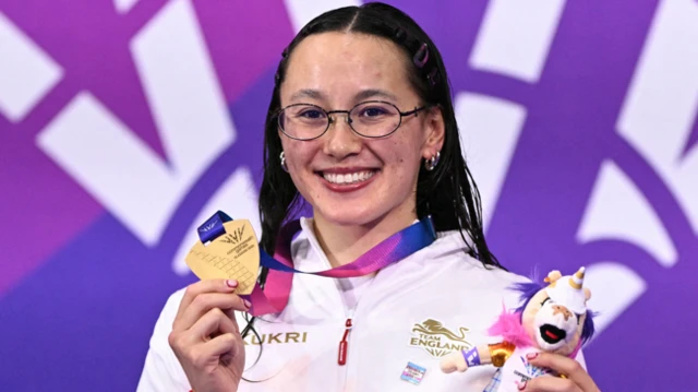 Alice Tai, wearing England's white swimming jacket, celebrates winning the women's 100m backstroke S9 by holding up her medal in her right hand