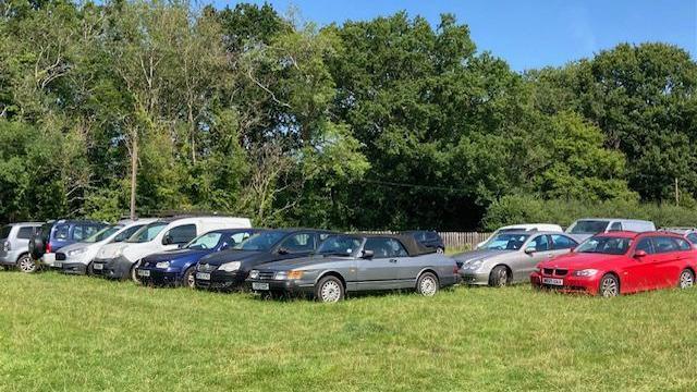 Rows of cars seen in a field, with trees in the background