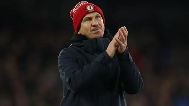 Gerhard Struber wearing a red Bristol City beanie hat and dark overcoat applauds supporters